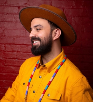A bearded man wearing a yellow shirt and a brown hat, smiling and looking to the side, with a beaded necklace, standing against a red brick wall.