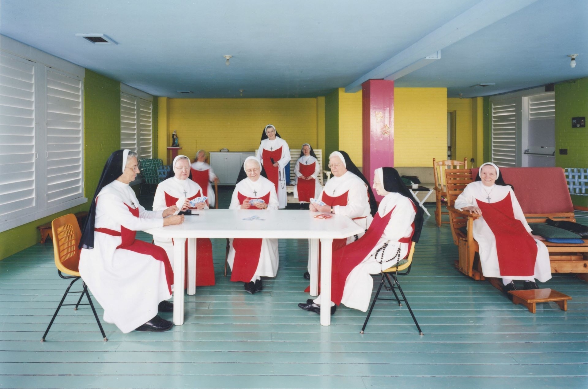 A group of nuns in white and red habits, sitting around a table in a brightly colored room, engaging in activities together.
