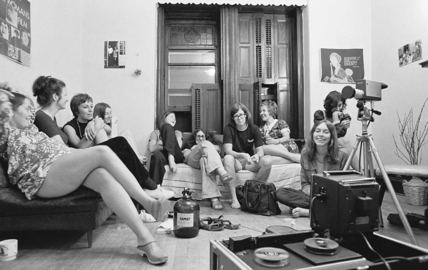 A black and white photo of a group of women sitting and laughing together in a room with a video camera setup, capturing a casual and joyful moment.