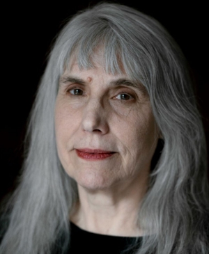 A headshot of an woman with long, silver hair, wearing a black top and looking directly at the camera.