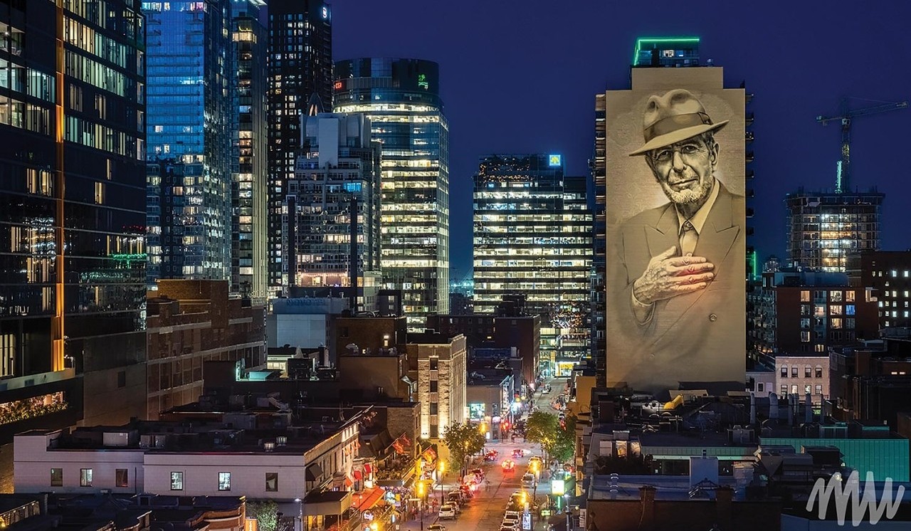 A vibrant nighttime view of downtown Montreal with a prominent 21-story mural of Leonard Cohen, created by artists El Mac and Gene Pendon, illuminated on Crescent Street.