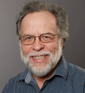 Headshot of a man with dark grey hair and white beard. He is wearing a blue polo shirt and glasses, and is smiling in front of a grey background