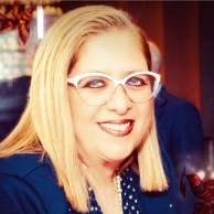 A woman with blonde hair, glasses and a blue jackets smiles for a portrait.