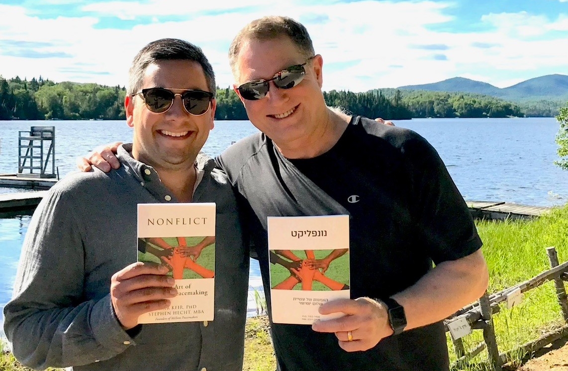 Image of two men smiling outdoors, in front of a lake. They are each holding a book and are wearing sunglasses.