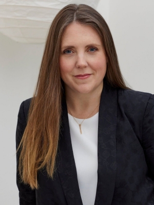 She wears a white top, black blazer and a thin gold chain necklace. Her hair is straight, long, centre-parted, and dark blonde. She has blue eyes.