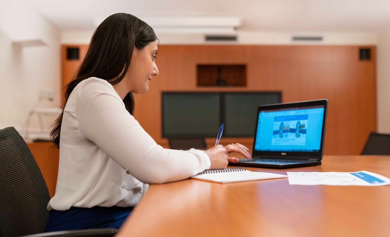 A woman with dark long hair is wearing a white blouse and sitting at a boardroom table taking notes while looking at a laptop