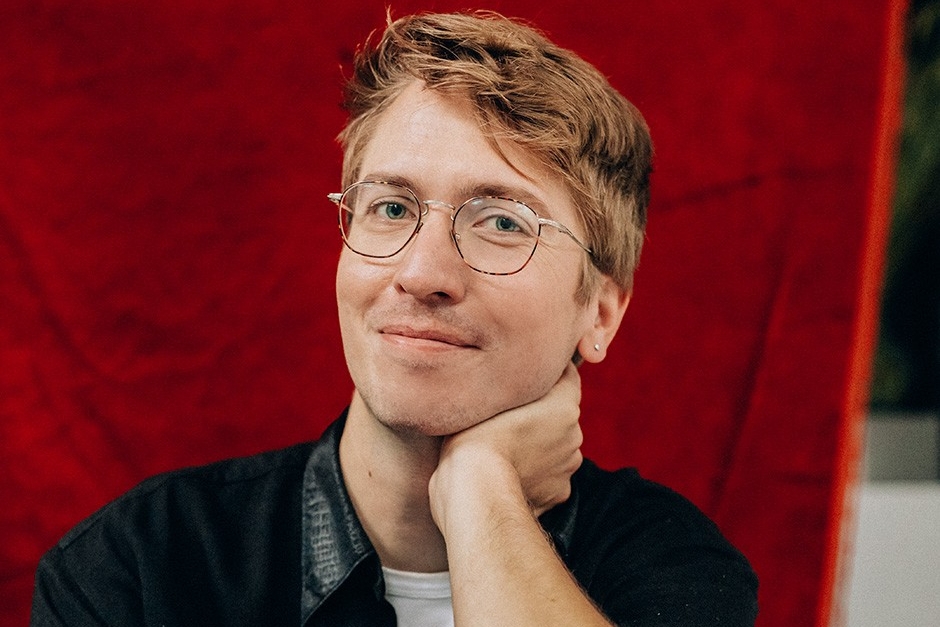 Devon Bate, who has blonde hair and wire-rimmed glasses, poses in front of a red background, resting his chin on his left hand