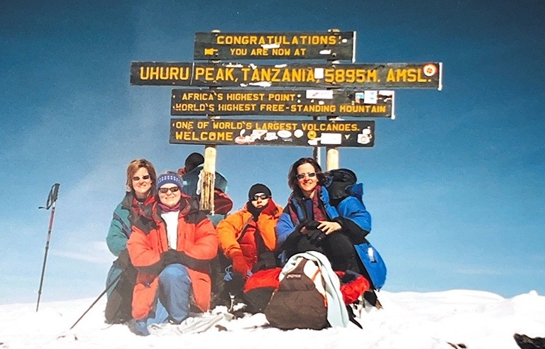 Four women in climbing gear pose in front of the sign at the top of Mount Kilimanjaro