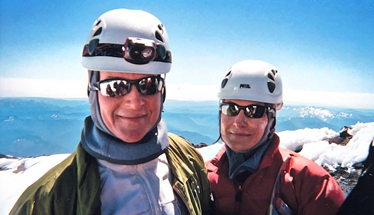 Two women pose in hiking helmets at the top of a mountain with snowy peaks behind them