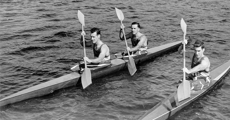 In a black and white image from 1960, three men are paddling down a waterway in narrow kayaks