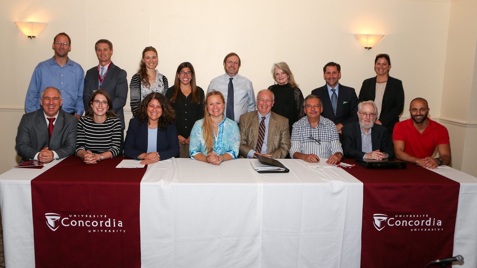 A row of people sit behind a long table with another row of people standing behind them