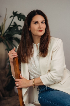 Woman sits on a wooden chair with a plant in the background. She has long brown hair and wears a white shirt, white blazer and blue jeans.