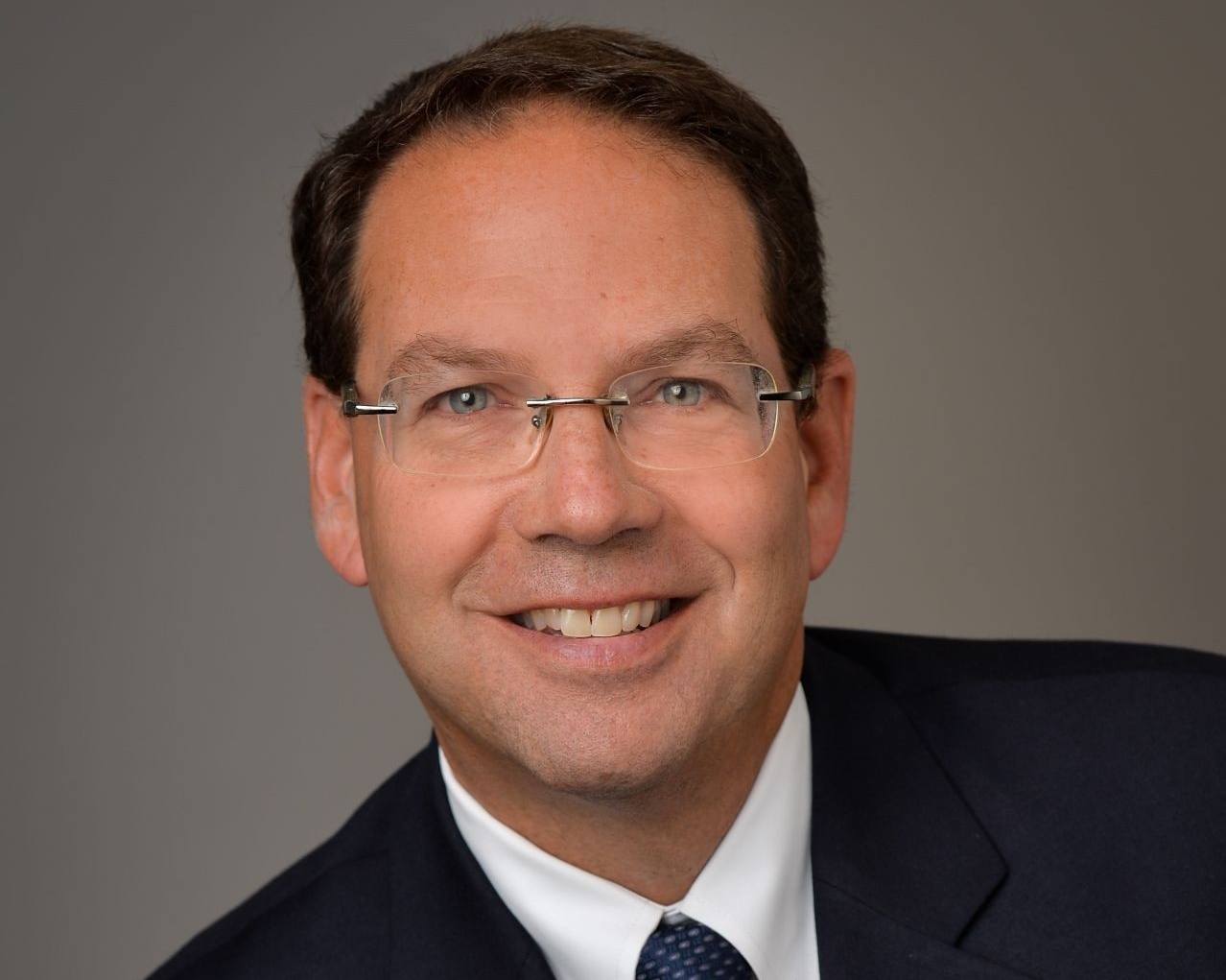 Man in a blue suit, white shirt, blue tie and glasses sit smiling for a portrait.