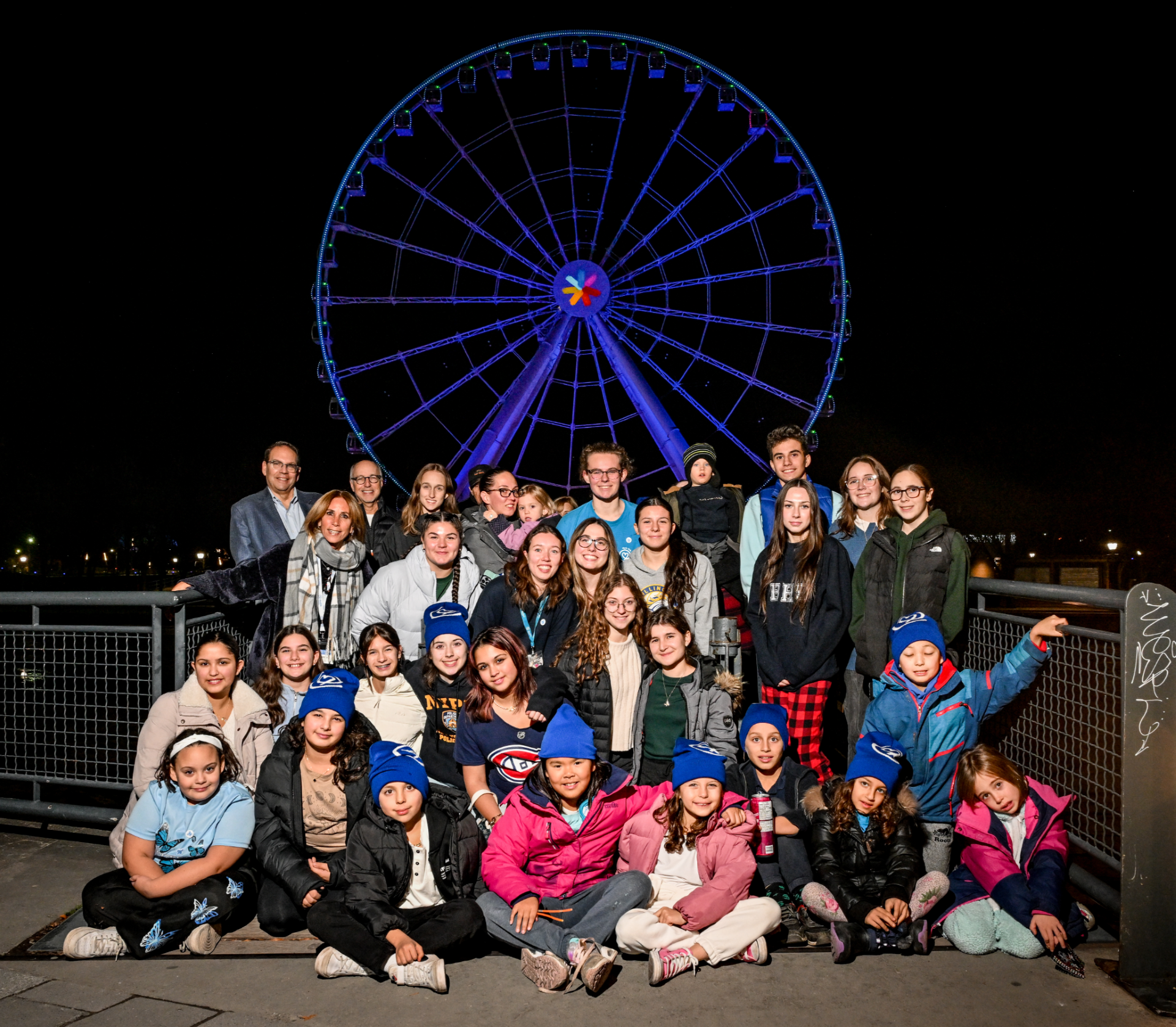 A group of children adults pose as a group in from of a large ferris wheel that is lit up in blue lights.