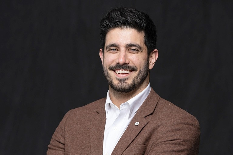 A young man with short dark hair wears a white shirt under a browm tweed jacket. He has a CFL pin on his lapel.