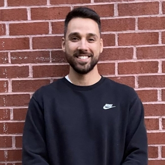 A bearded male with brown hair stands in front of a brick wall smiling for a portrait.