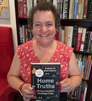 A woman with curly hair stands holding a book stands in front of a bookshelf smiling for the camera.