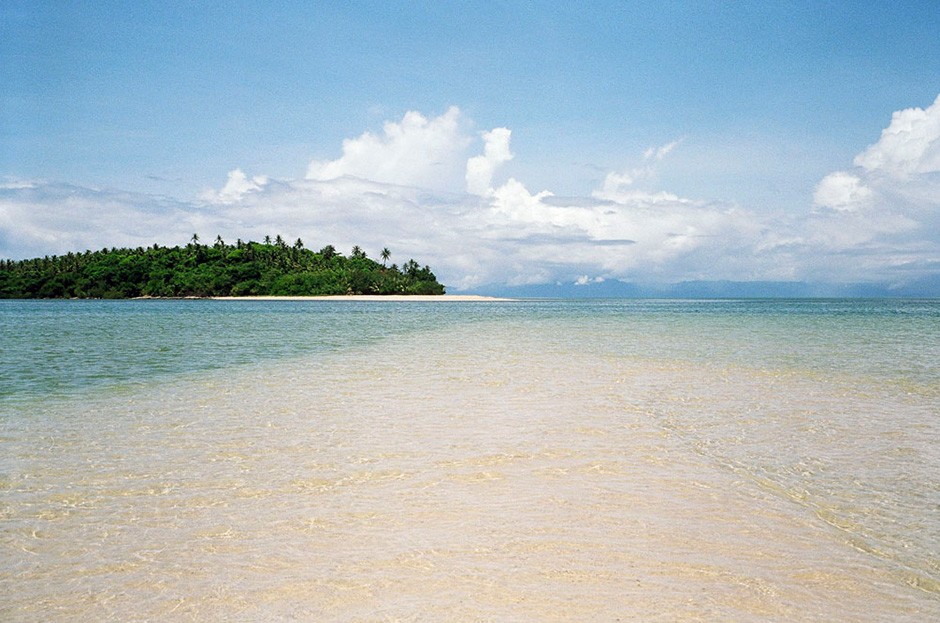 A sandy beach stretches out towards the horizon to meet a bright blue sky. Where they meet there is a small group of green trees to the left.