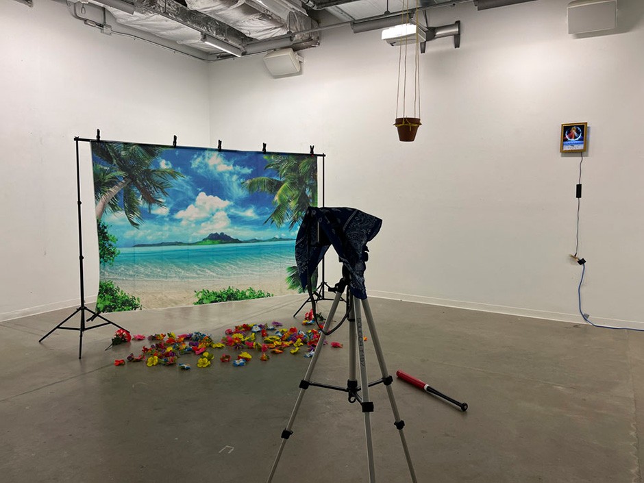 In the corner of a spare gallery space, a screen is set with an image of a tropical island scene. Multi-colored flowers are strewn across the floor below it. To the right of the screen a red and black baseball bat lies on the ground. A plant holder hangs from the ceiling and a clock with an illuminated face hangs on the wall to the right of it. In the foreground there is a camera on a tripod.