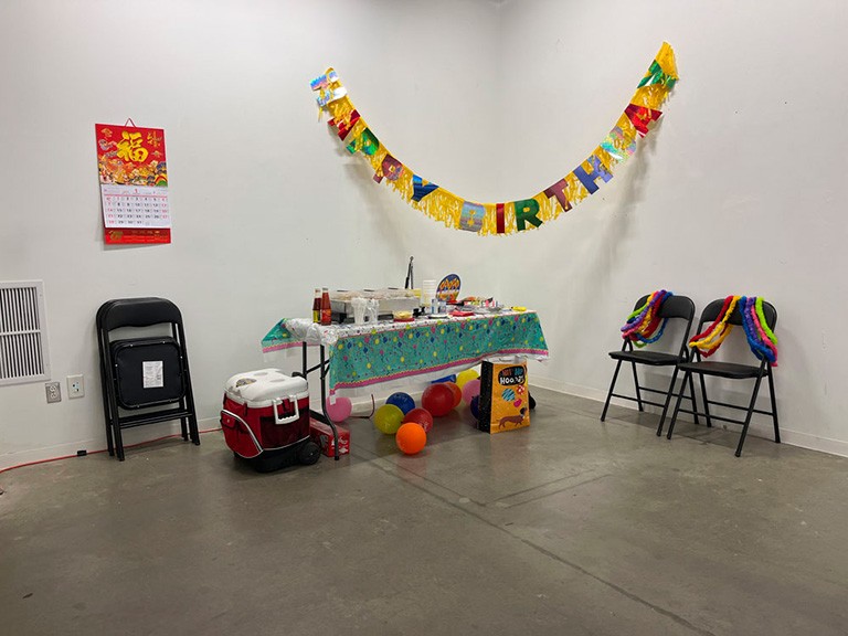 In the corner of a room there is a table set with party food and drinks. Above it is a streamer with Happy Birthday on it. To the left of the table, above two folded chairs that are leaning against the wall is a calendar. Under the table are ballons, a red cooler and a gift bag. To the right of the table sit two chairs with rainbow streamers draped over them.