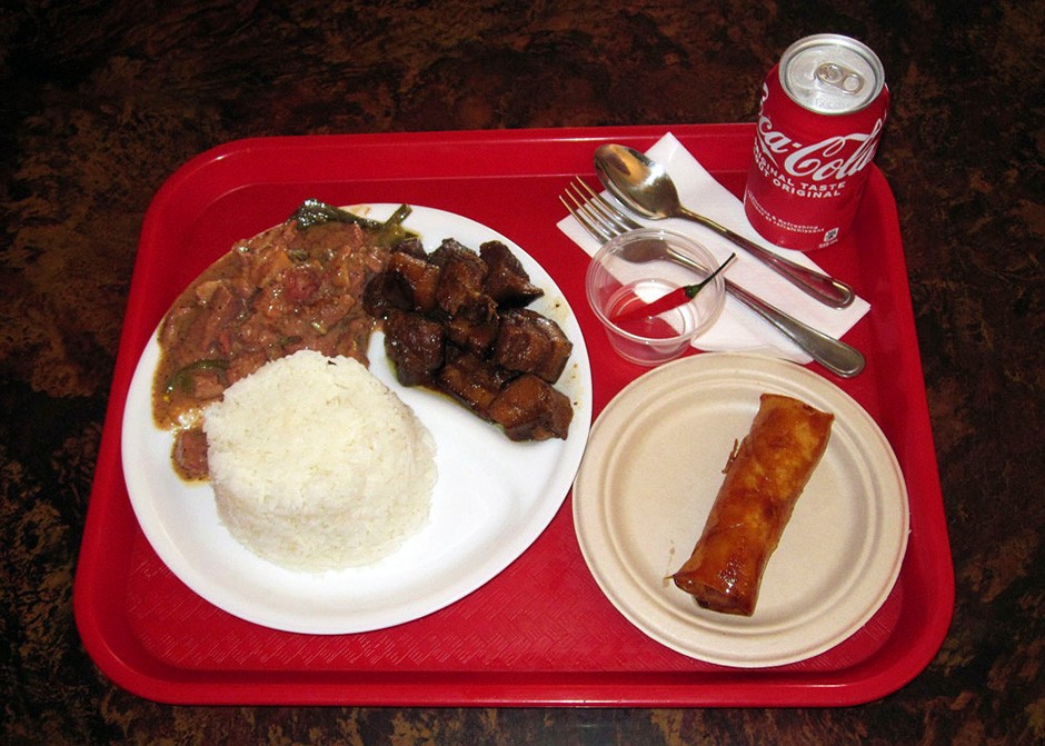 An overhead view of a red tray with two paper plates on it. The larger plate holds white rice, stew and potatoes; the smaller holds turon: ripened bananas are wrapped in spring roll wrappers. Also on the tray are a can of Coca-Cola, a fork and spoon on a white napkin and a small plastic container with a red pepper in it.