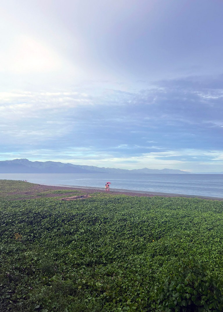 The bottom half of this vertical photo is green grass stretching up towards a shoreline where three small figures stand under an umbrella. The water is grey-blue and beyond it are mountains in the background, almost the same colour as the water. Clouds hang in the sky and take up the other hal of the photo.