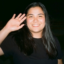 In front of a black background, Tyra Maria has long dark hair and wears a black t-shirt. She holds her hand up next to her face as if she is waving hello to you.