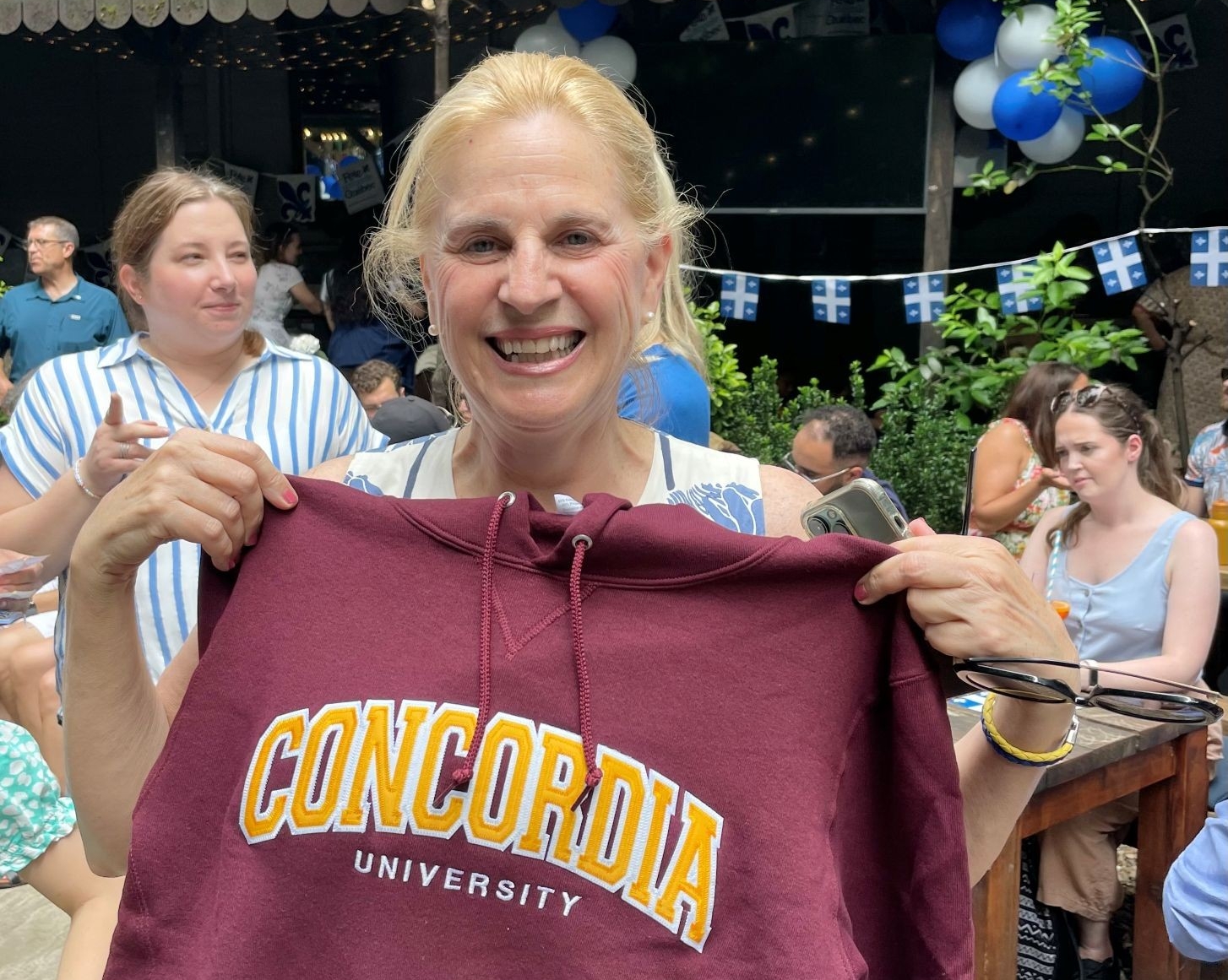  A woman holding a maroon Concordia University hoodie, smiling joyfully at an outdoor gathering with Quebec-themed decorations and other people in the background.