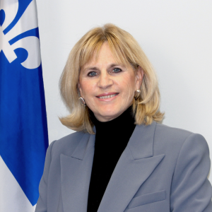 A professional portrait of a woman wearing a gray blazer and black turtleneck, standing in front of the Quebec flag, smiling confidently.