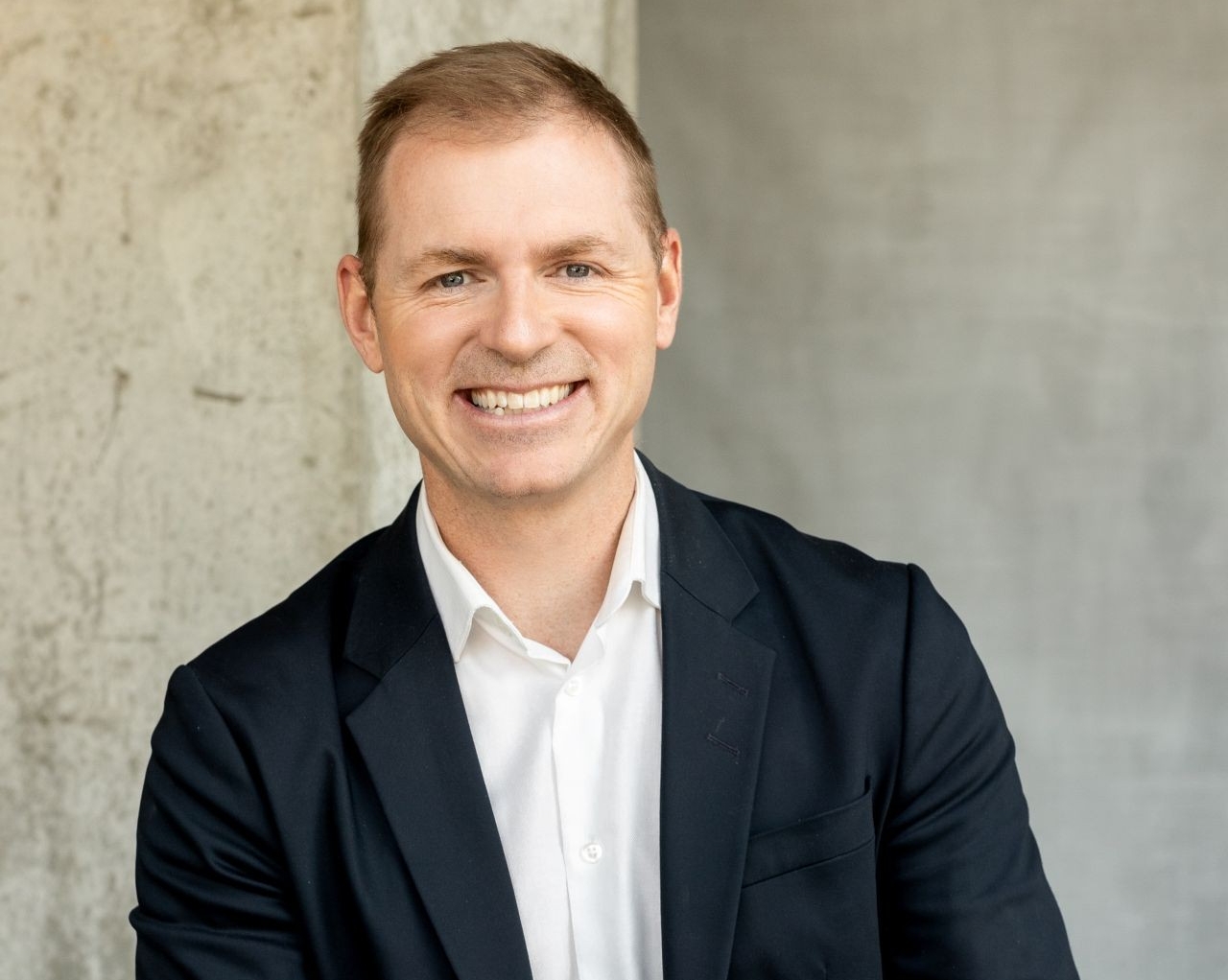 A male wearing a navy blue blazer and white collared shirt stands smiling.