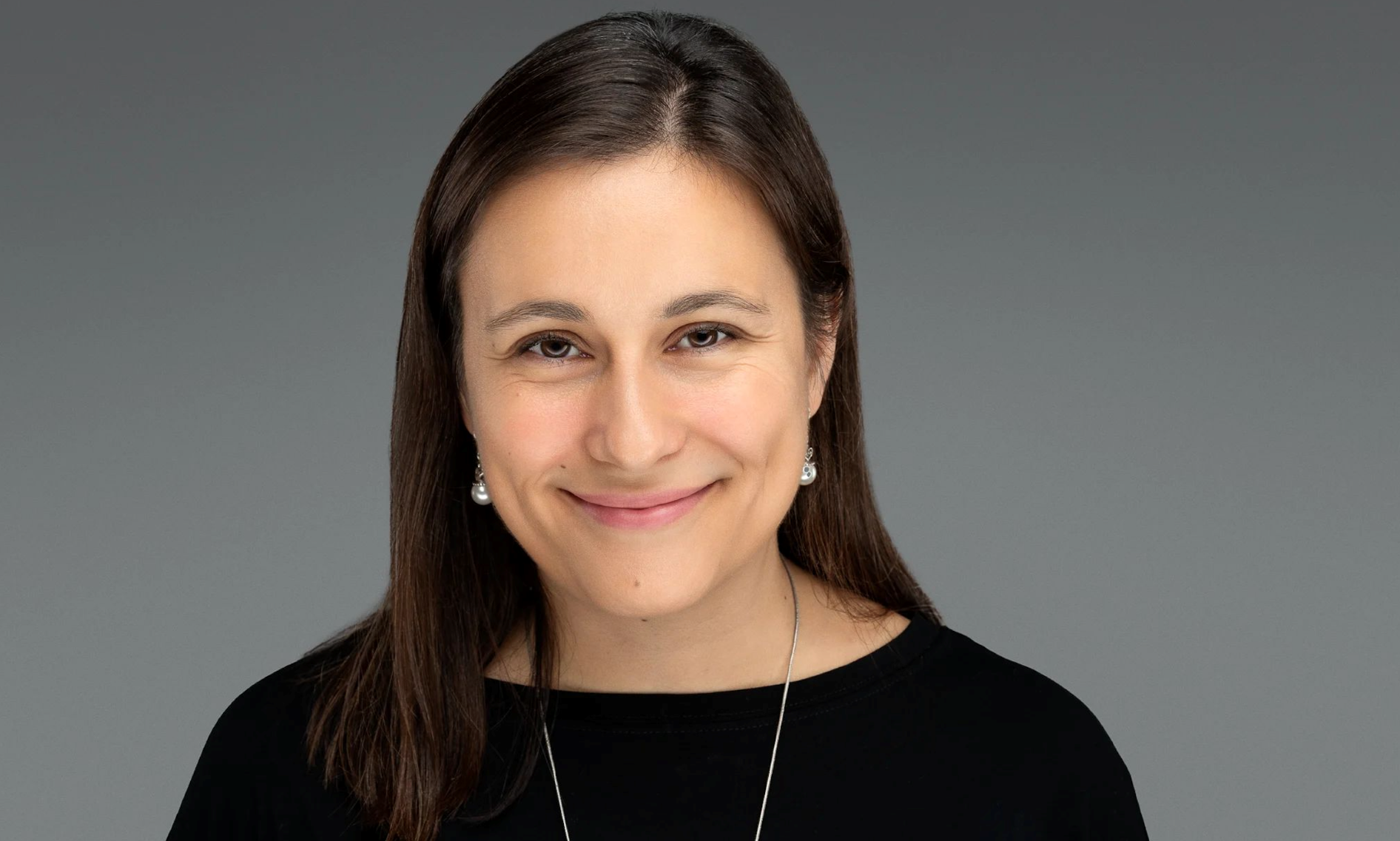 portrait of a smiling woman with long, dark hair, wearing a black top in front of a gray background