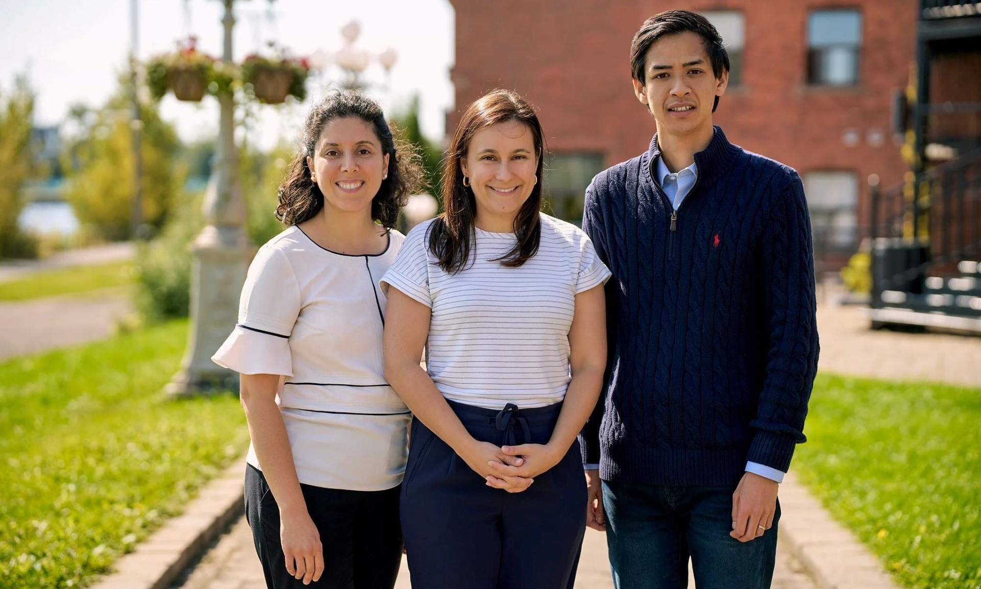 two dark-haired women wearing dark pants and short-sleeved white tops stand next to a dark-haired man wearing a navy sweater. A red-brick building and grass are in the background. 