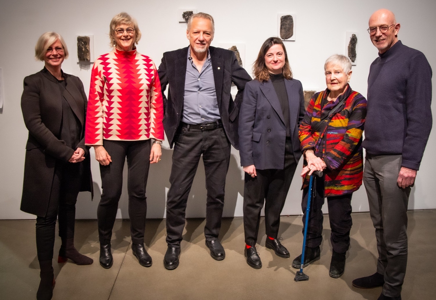 Six people stand next to each other in front of a white wall with art displayed on it