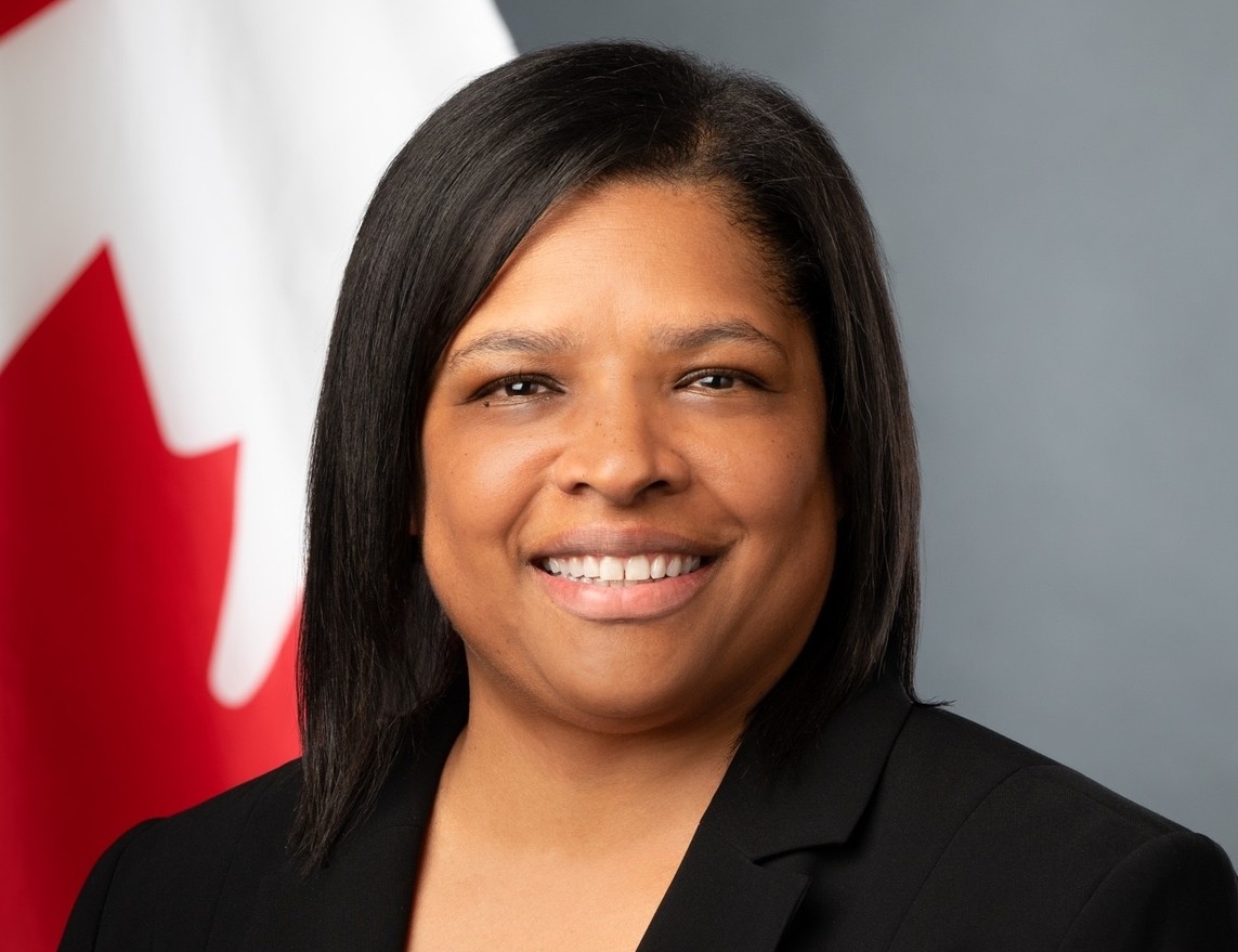 A woman with dark hair poses in front of a Canada flag. She is wearing a dark blazer over a red shirt 