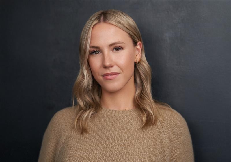 A woman with shoulder length blonde hair and beige sweater stands in front of a dark grey backdrop