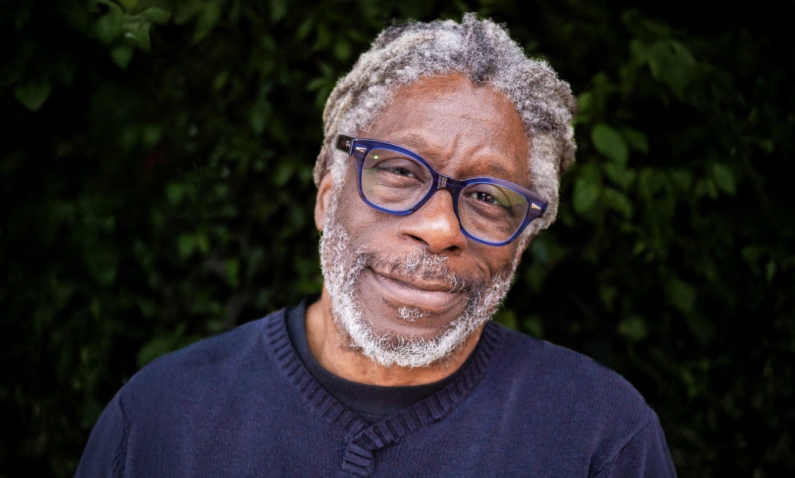 A man with grey hair and beard is wearing blue-framed glasses and wearing a navy blue sweater. He is standing in front of dark foliage