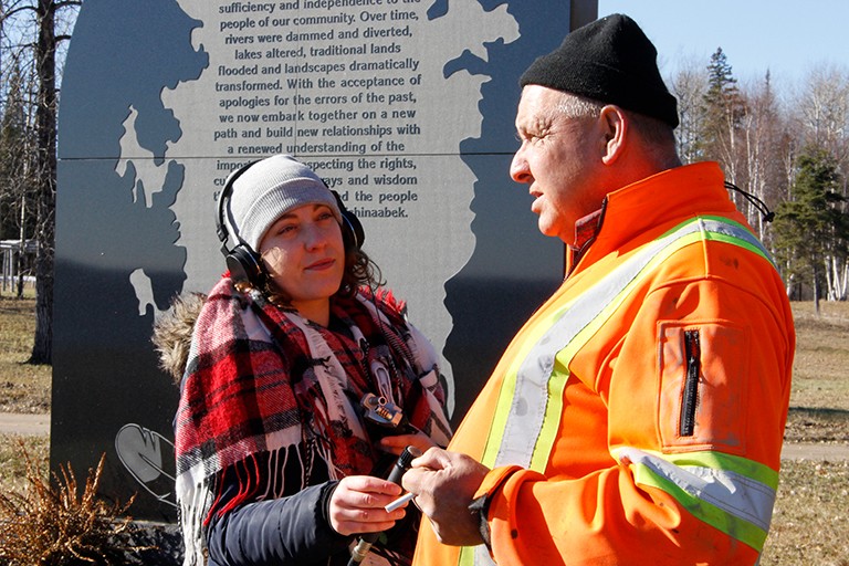 Une jeune femme portant un bonnet d'hiver, des écouteurs et une écharpe d'hiver, interviewant un homme plus âgé portant une salopette de chantier orange.