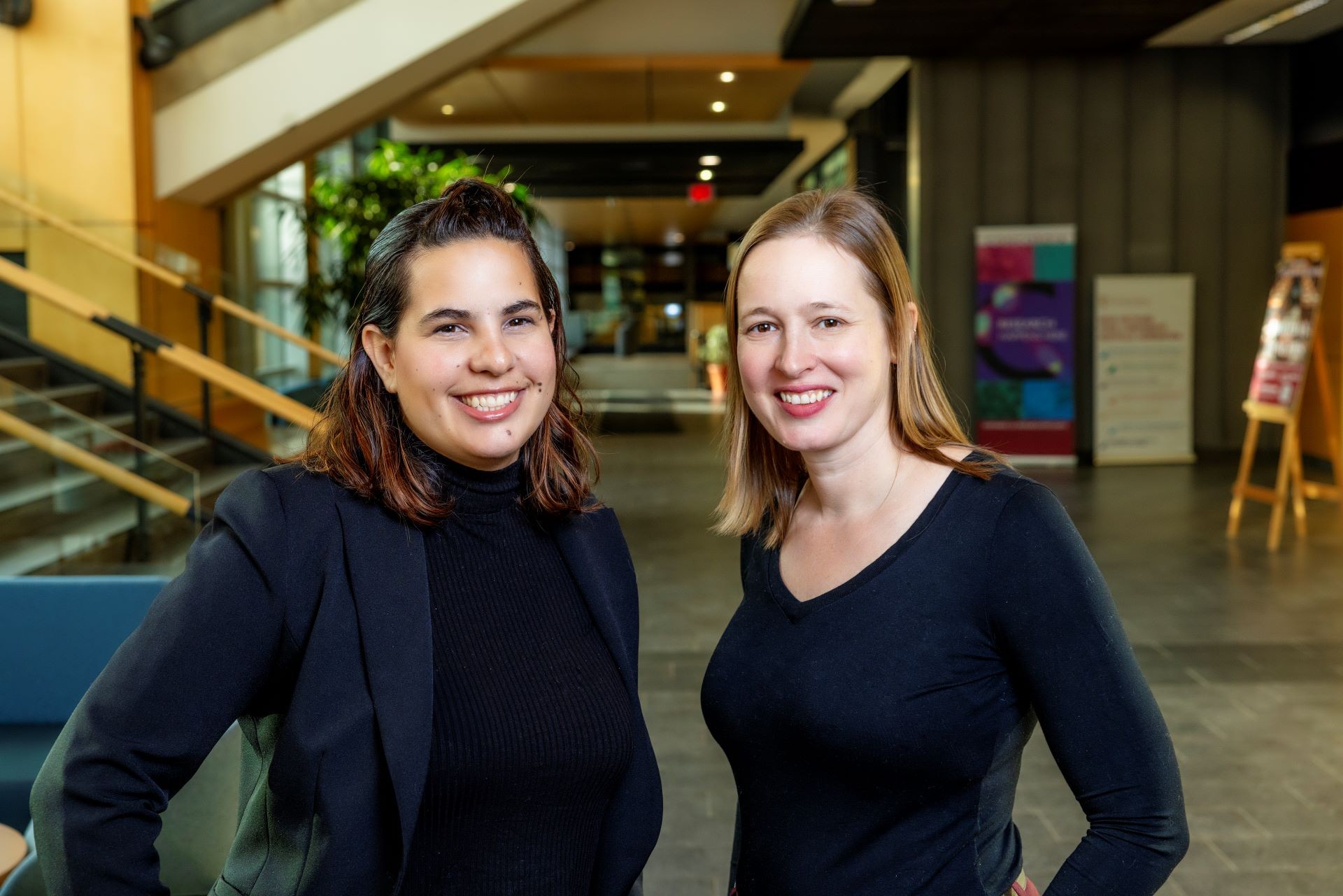 Two women dressed in dark clothes smile in a university corridor