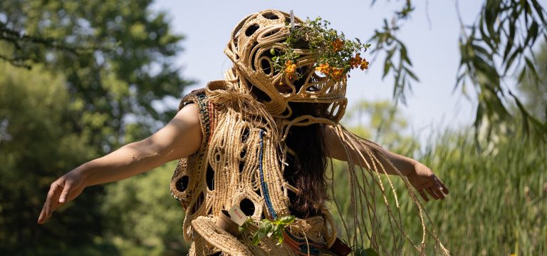 Une figure de femme en plein air, les bras tendus, portant un costume sculptural fait de fibres de corde et de fleurs.