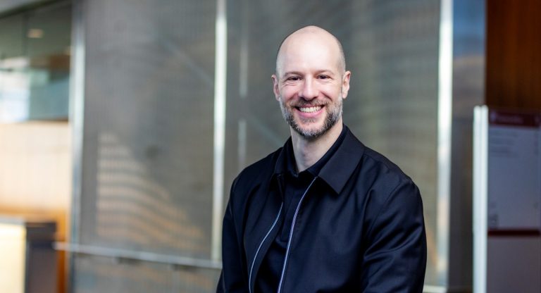 A smiling bearded man wears a black jacket and shirt