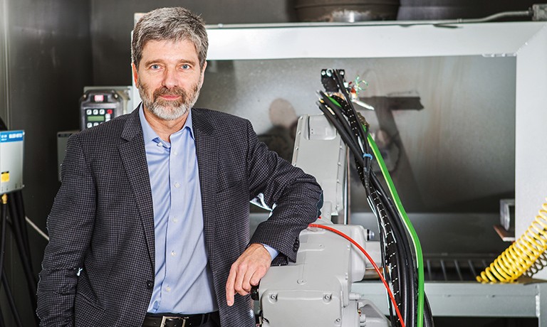 Slightly smiling man with grey hair and beard, wearing a suit and standing in an engineering mechanical lab.