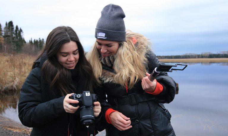 A young smiling woman with long, dark hair, wearing a black winter jacket, showing a shot on her camera to an older smiling woman with blonde hair wearing a black winter jacket and a grey beanie hat.
