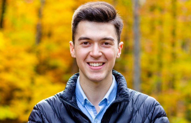 A smiling young man with brown hair, wearing a blue puffy jacket, with a blue collared shirt underneath, in front of a backdrop of fall foliage
