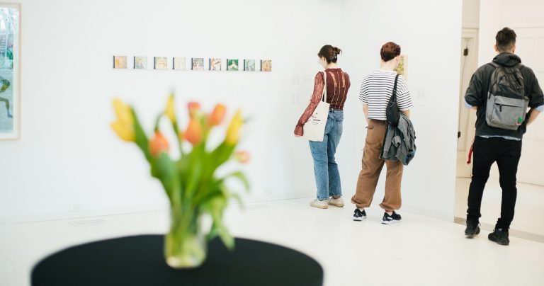 Three people in a white exhibition space, with a table and flowers in the foreground.