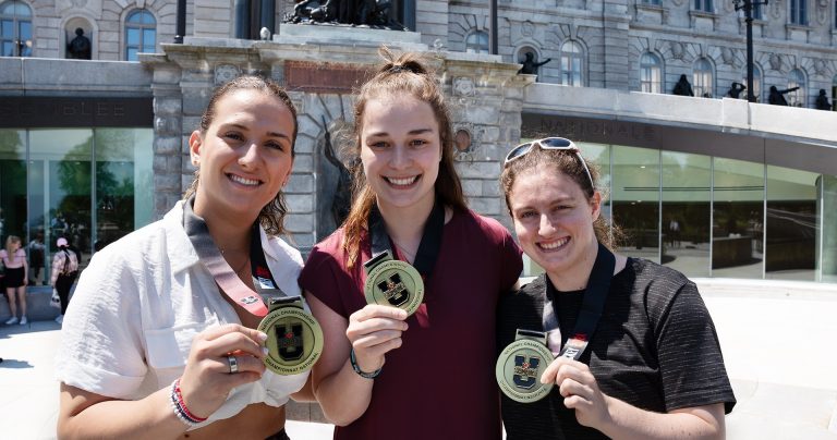 Three young women standing outside in front of parliament buildings, holding up gold medals and smiling.