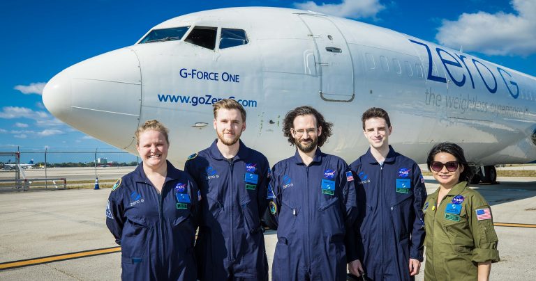 A group of smiling men and women in jumpsuits standing in front of an aeroplane with the words "G-Force-One" on the side.