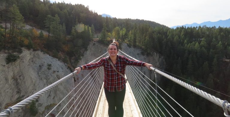 A smiling young woman wearing a plaid shirt and black leggings standing on a long swing bridge over a ravine.
