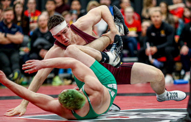 Two young men, one in burgundy and the other in green, wresting in front of an audience at a competition 