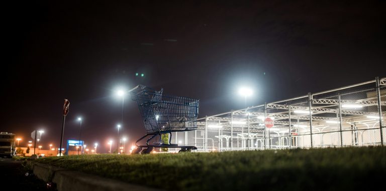 A night shot with a shopping cart in the foreground and a lit-up shopping mall in the background.