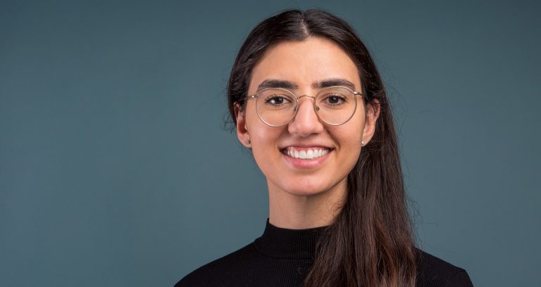 Smiling young woman with long, dark hair, wire-rimmed glasses and a black turtleneck sweater.